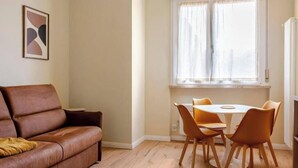 Ein Wohnzimmer mit braunem Ledersofa, einem weißen runden Tisch mit vier Stühlen, einem Fenster mit Vorhängen und einem gerahmten abstrakten Kunstwerk an der Wand.