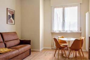 Ein Wohnzimmer mit braunem Ledersofa, einem weißen runden Tisch mit vier Stühlen, einem Fenster mit Vorhängen und einem gerahmten abstrakten Kunstwerk an der Wand.