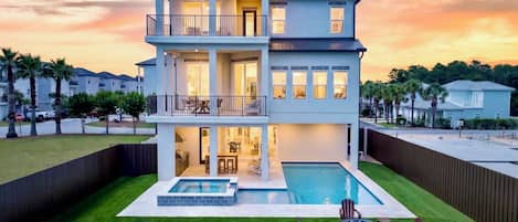 A two-story house with a swimming pool, outdoor seating, and a view of the sunset.