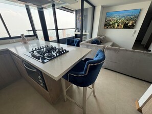 A modern kitchen with a gas stove, a dining area with a white table and blue chairs, a sofa, and a large window offering a city view. - Penthouse in the best view of Medellin (Medellín)