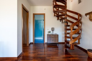 Un escalier en colimaçon se trouve dans un couloir dont le sol est en bois, et une porte donne accès à une pièce dont le sol est recouvert de carreaux bleus.