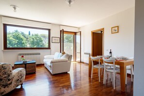 A spacious living area with a large window, a white sofa, a dining table with chairs, and a view of trees outside. - Apartment 'Posta Del Lago' with Lake View, Private Terrace and Wi-Fi (Passignano sul Trasimeno)