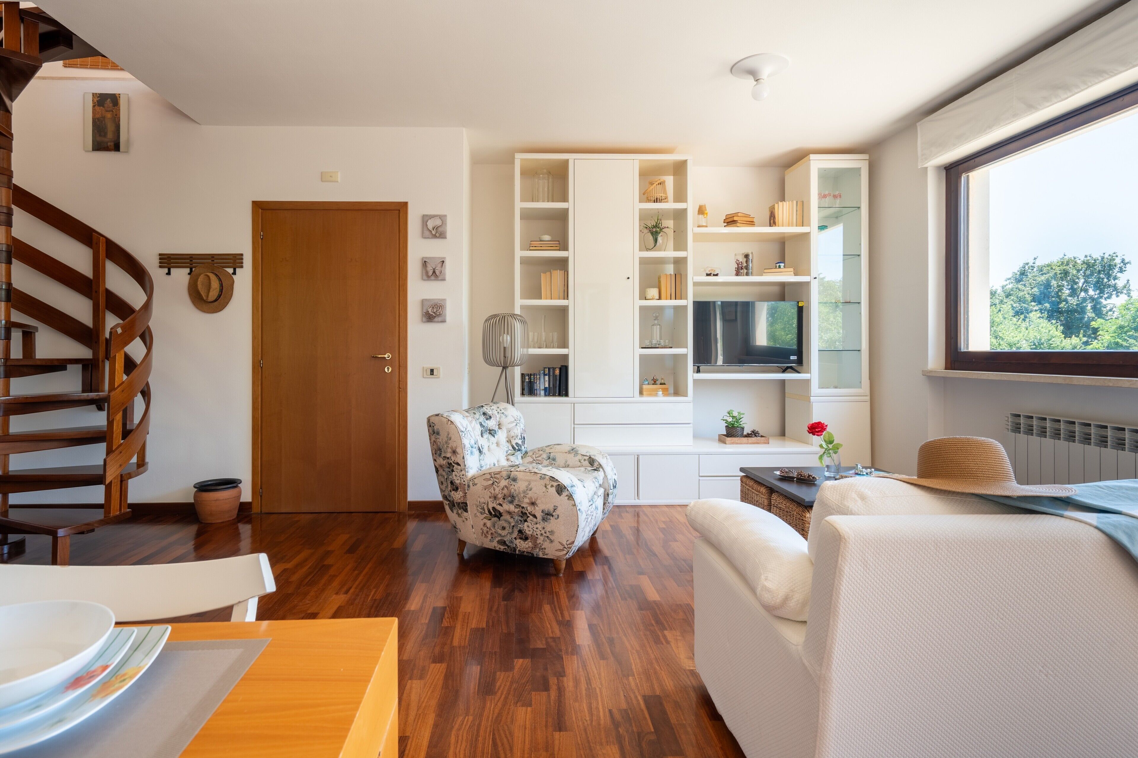 A modern living room with a wooden staircase, a floral armchair, a sofa, and a built-in TV shelf.