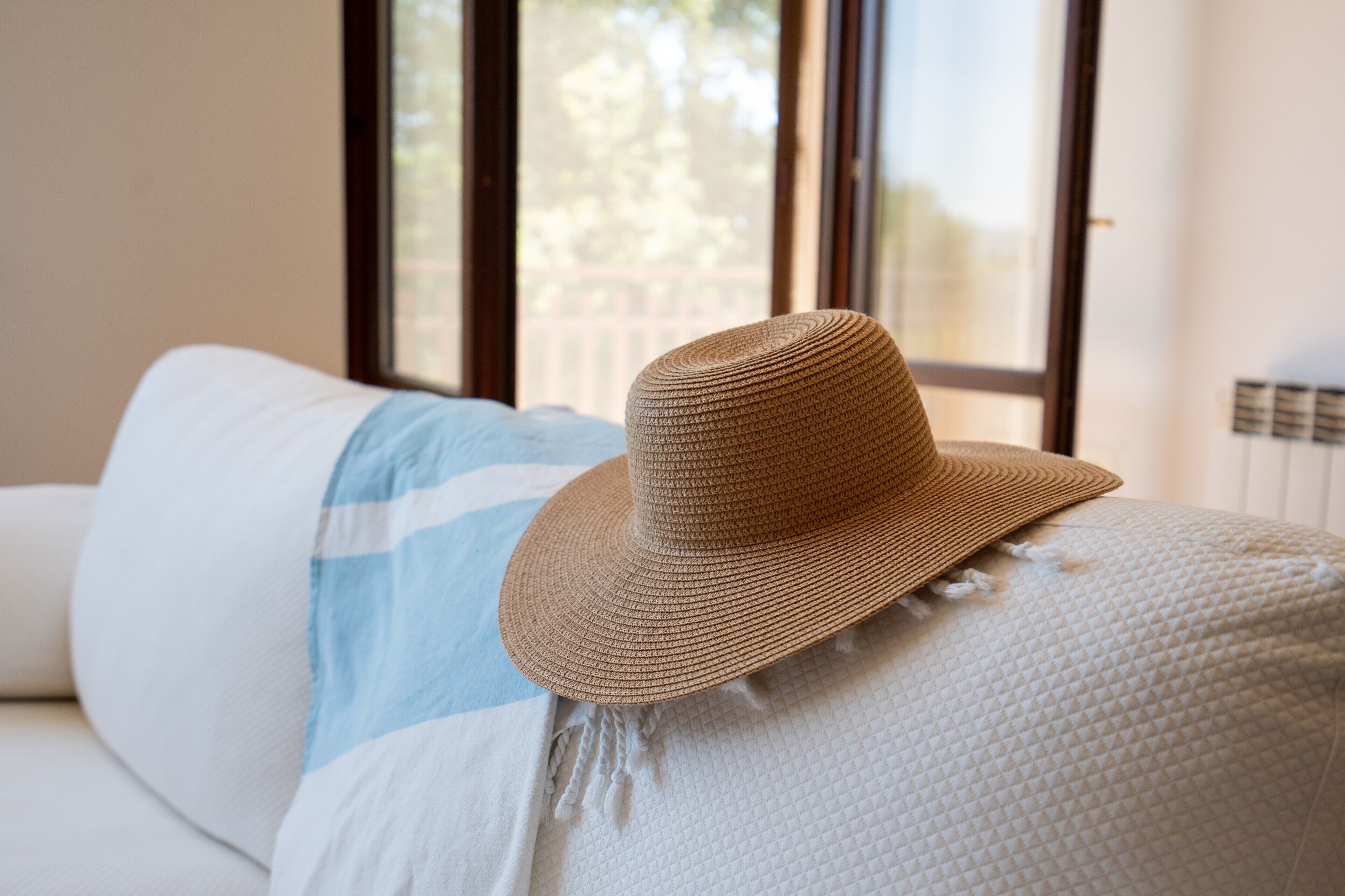 A room with a large window, a white sofa, and a woven hat on the armrest.
