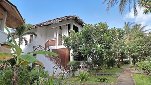 Un edificio blanco con techo de paja, una escalera de madera y un sendero de piedra rodeado de vegetación tropical.