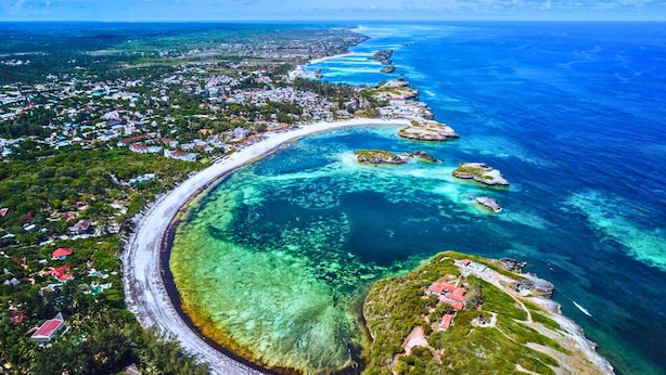 Vista aérea de un pueblo costero con una carretera serpenteante, aguas azules cristalinas y edificios dispersos.