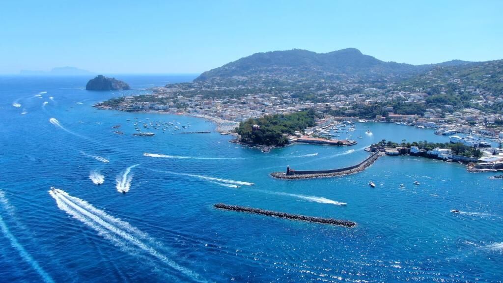 Vista aérea de un pueblo costero con un puerto deportivo, barcos y montañas al fondo.