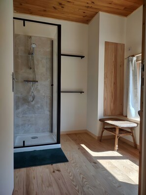 A modern bathroom with a glass-enclosed shower, wooden flooring, a wicker chair, and a wooden ceiling.