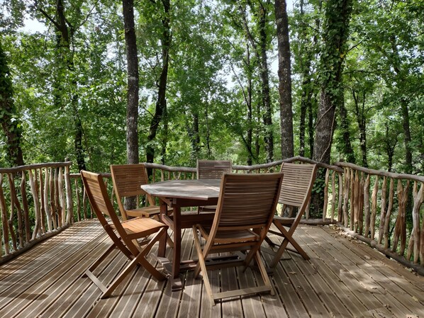 A wooden deck with a table and chairs surrounded by a wooden railing, set in a forested area.