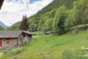 Edifici de pedra rústic amb teulada inclinada, envoltat d'herba verda i arbres frondosos.