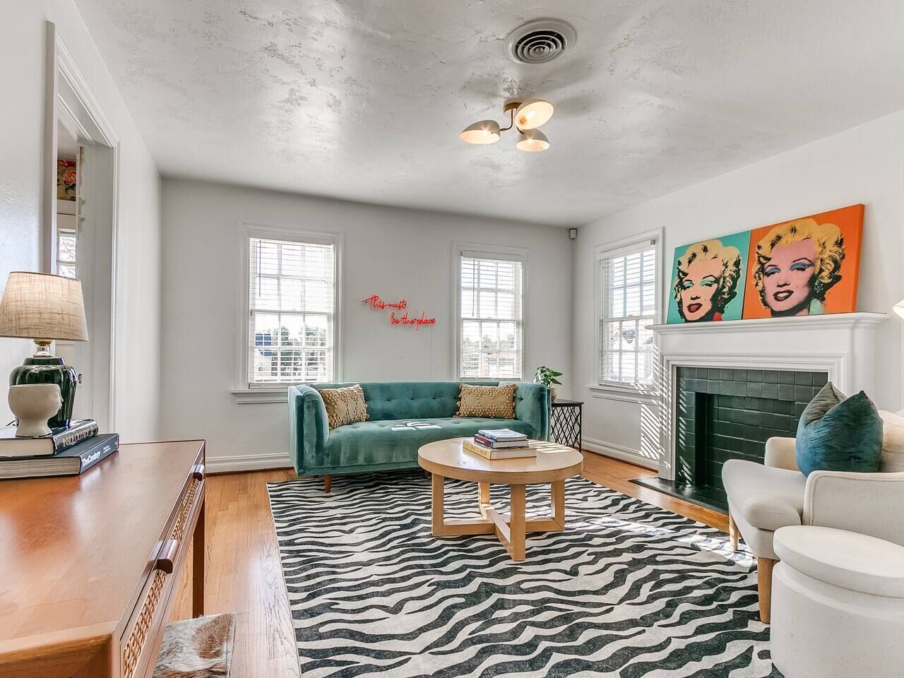 A living room with a teal sofa, a round wooden coffee table, a zebra-patterned rug, a white fireplace, and a painting of Marilyn Monroe.