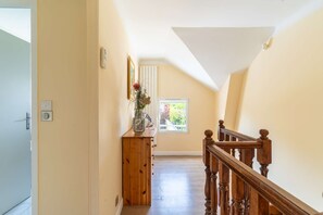 Ein Flur mit einer Holztreppe, einem Fenster mit Blick auf Bäume und einer Vase mit Blumen auf einer Holzkommode.