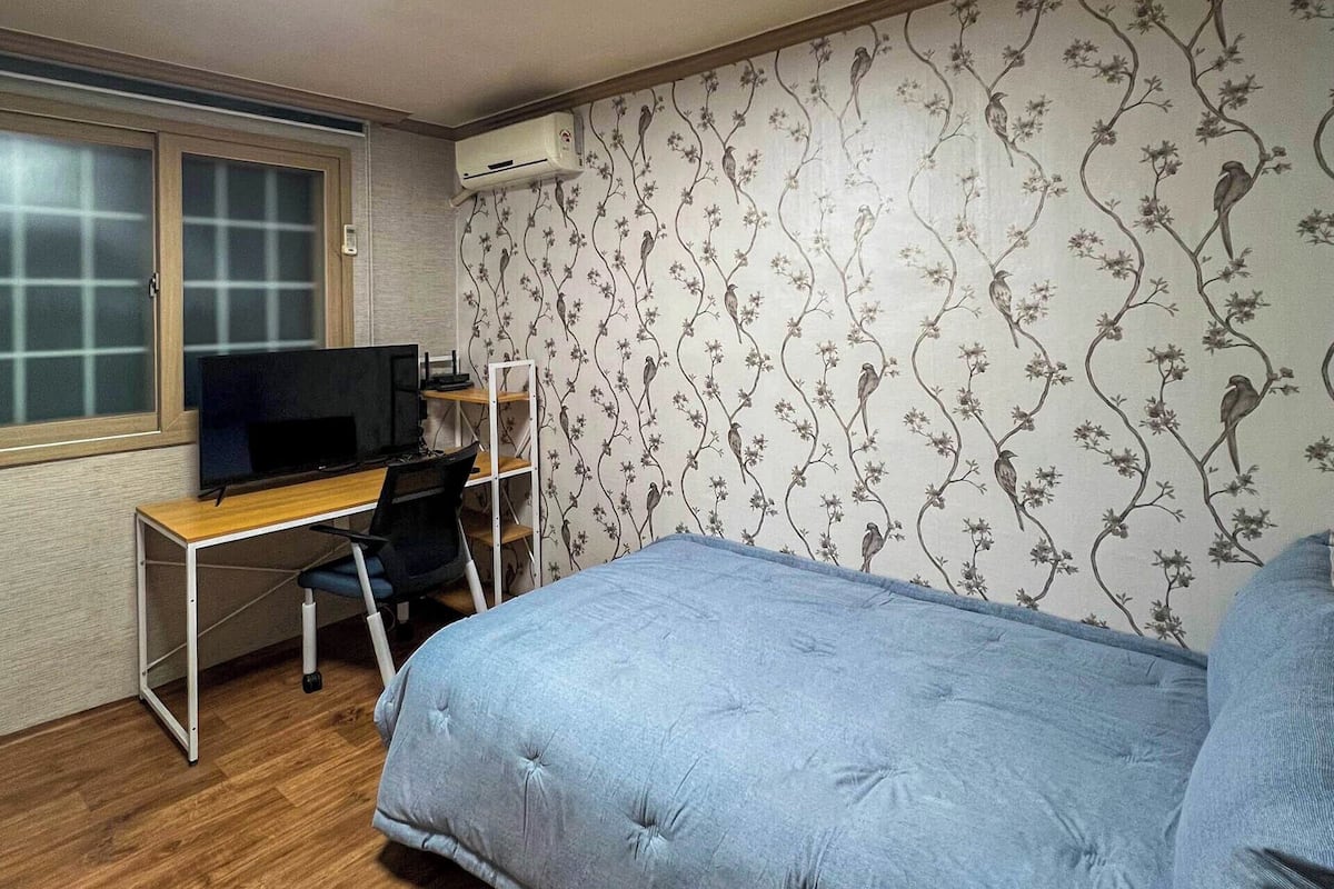 A bedroom with a bed, a desk with a chair, a TV, and a window.