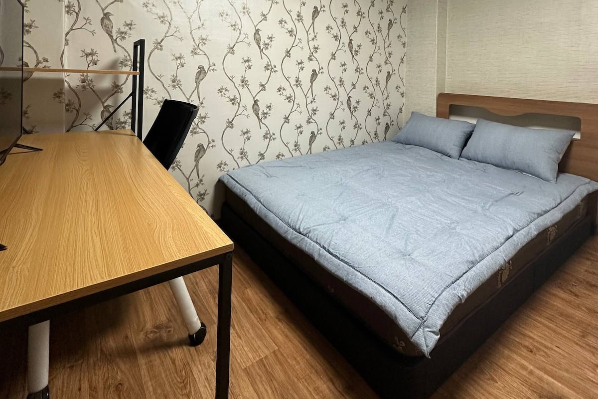 A bedroom with a wooden desk, a bed with a grey mattress, and a floral wallpaper.