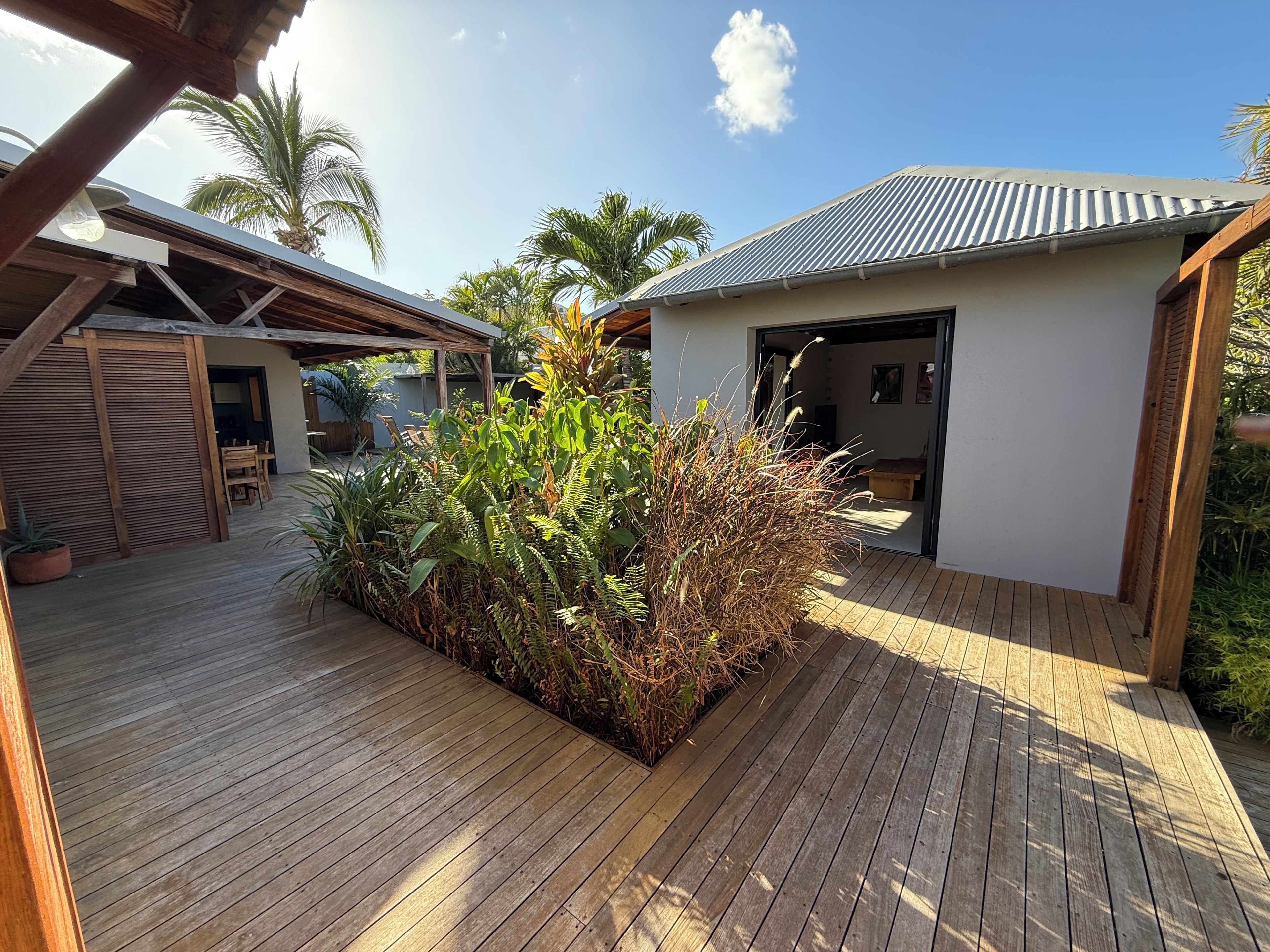 A wooden deck with a planter filled with tropical plants, a covered outdoor seating area, and a building with a corrugated metal roof.