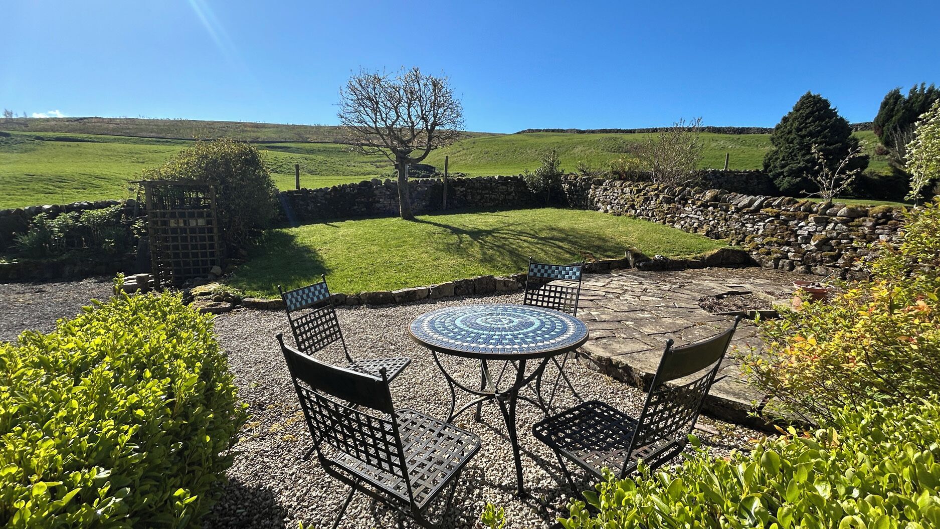 Une terrasse agrémentée d’une table et de chaises en mosaïque, entourée de verdure et de murs en pierre.