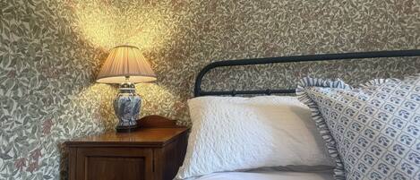 A bedroom with a patterned wallpaper, a bedside lamp, a wooden nightstand, and a bed with patterned bedding.