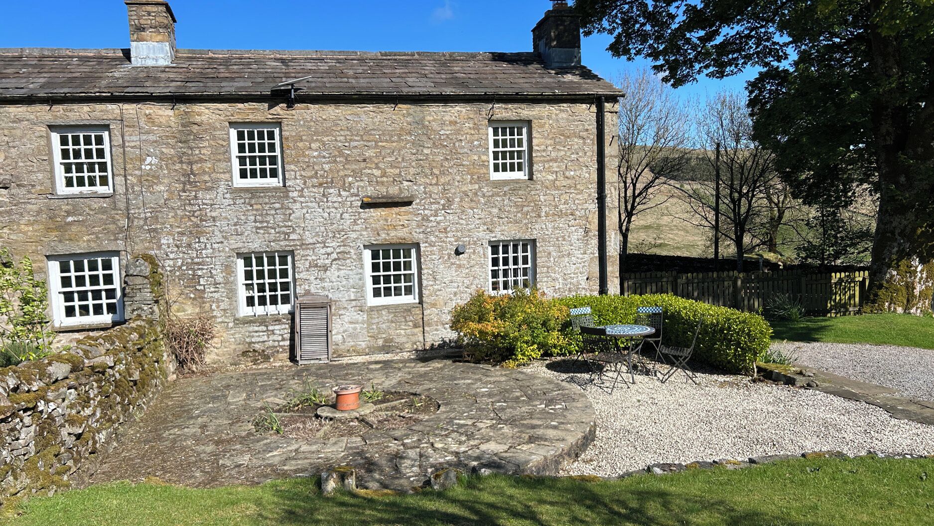 A stone building with multiple windows, a gravel driveway, and a small garden area with a table and chairs.