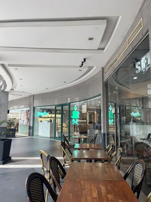 A modern indoor dining area with a long wooden table and wicker chairs, a glass storefront with a neon sign, and a curved ceiling with recessed lighting. - NEW! Near Venice Grand Canal Mall, BGC & Airport, w/ SAUNA, POOL, GYM & NETFLIX (Taguig)