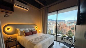 Een hotelkamer met een groot bed, een nachtkastje met een vaas, een aan de muur bevestigde televisie, en een balkon met uitzicht over de stad.