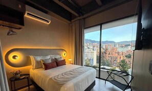 A hotel room with a large bed, a bedside table with a vase, a TV mounted on the wall, and a balcony with a city view. - Spectacular apartment in the town, surrounded by nature, parks and restaurants. (Medellín)
