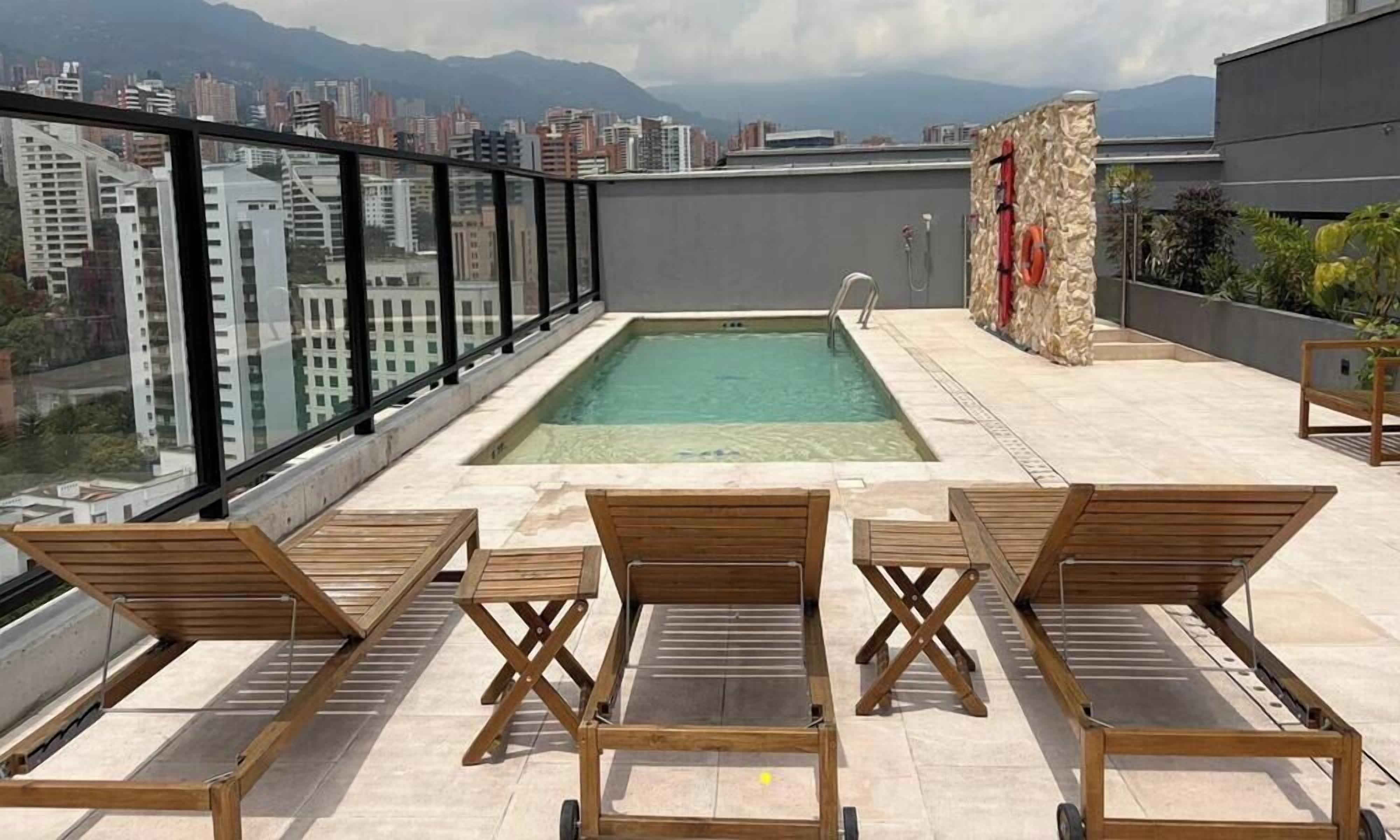 A rooftop pool with a city view, wooden loungers, and a stone wall with a red pipe.