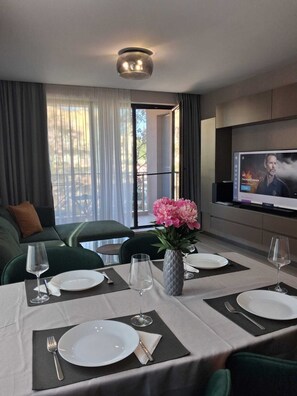 Ein Hotelzimmer mit Essbereich, einer Couch, einem Fernseher und einem Balkon mit Vorhängen.