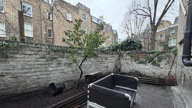 Un patio con un muro de ladrillo, un árbol pequeño y dos botes de metal negros.