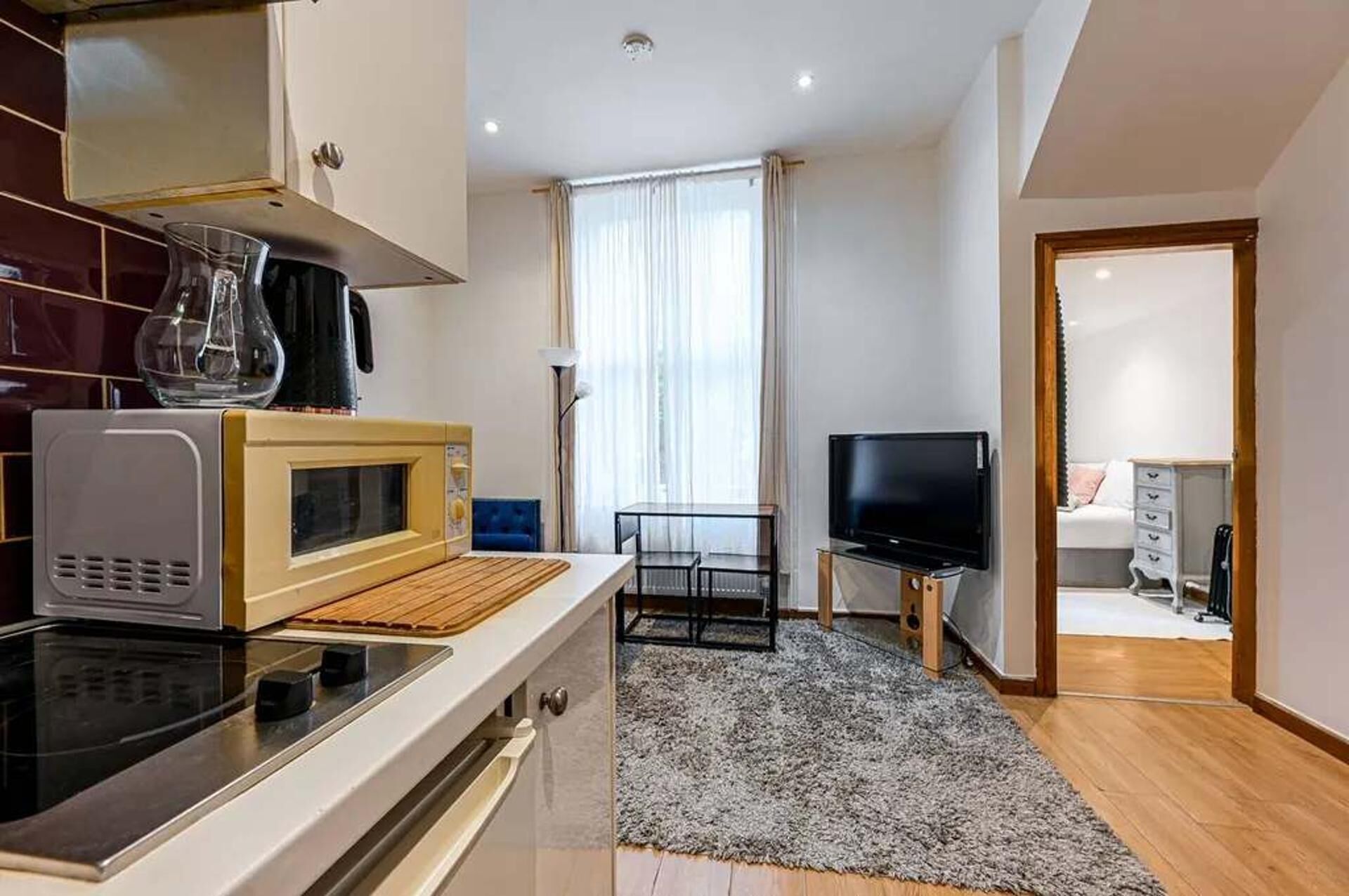 A compact kitchen with a microwave, stove, and a coffee maker. There is a TV and a small table with a chair. The room has a large window with curtains.