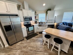 A compact kitchen with a refrigerator, stove, microwave, and sink. There are two stools at a wooden table. - The Bungalow – Escape, Explore, Enjoy (Garden City)
