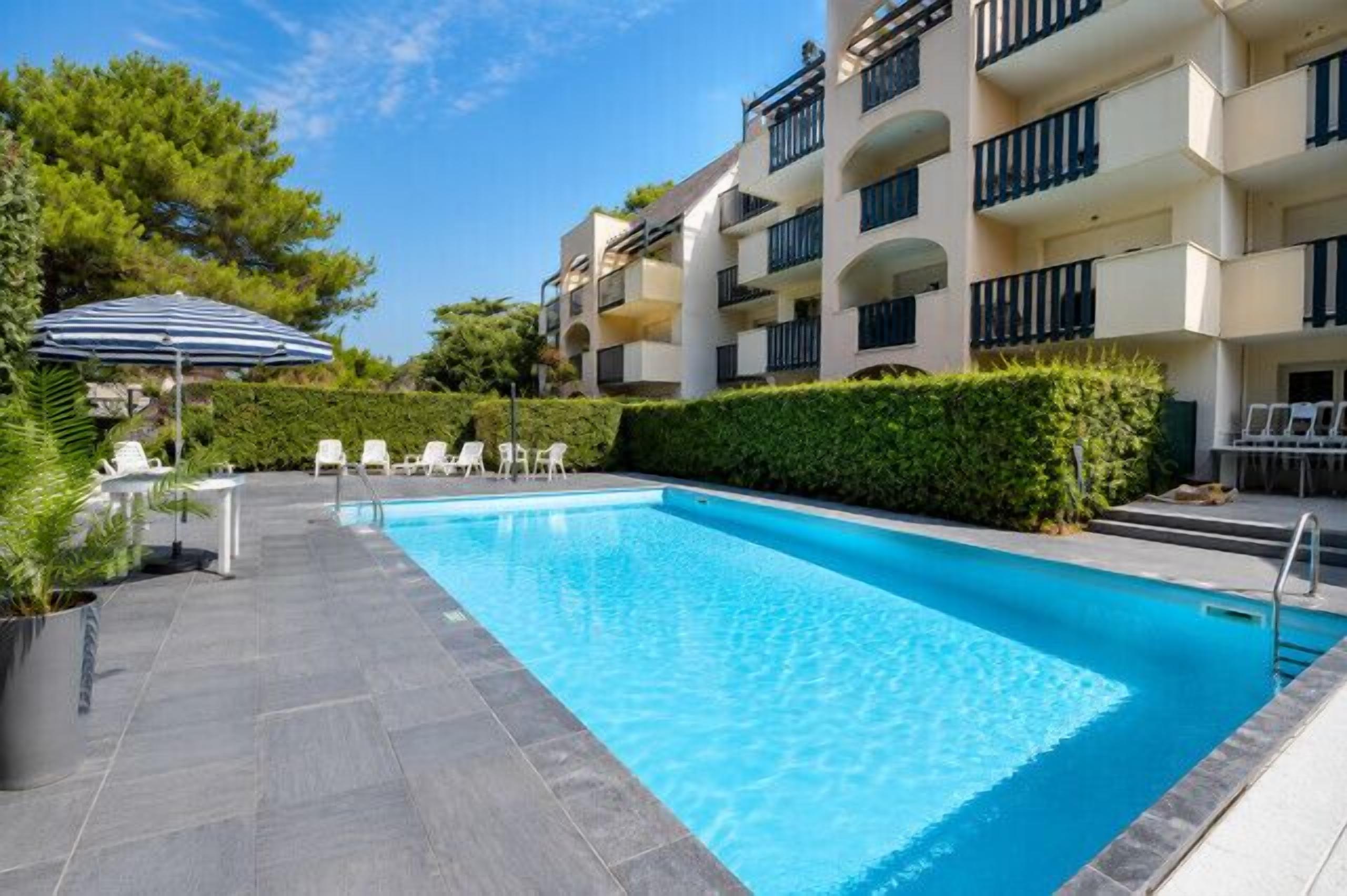 Una piscina con lettini, un ombrellone e un edificio a più piani con balconi.