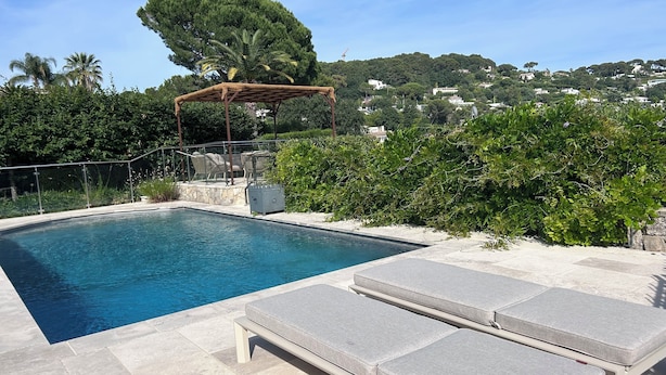Piscina com espreguiçadeiras, pérgola e vista para uma colina com árvores e casas.