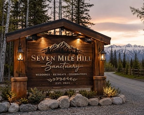 A wooden sign for Seven Mile Hill Sanctuary with a mountain silhouette, established in 2023, and mentions weddings, retreats, and celebrations.