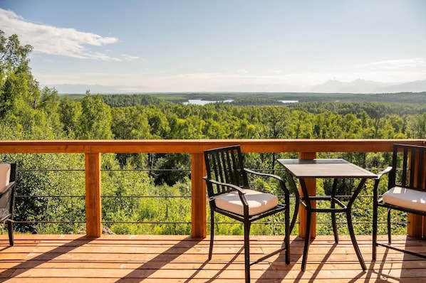 A wooden deck with a table and chairs overlooking a forested landscape with a lake and mountains in the distance.