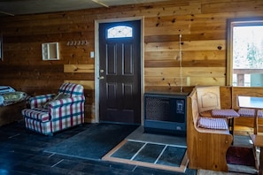 A cozy cabin interior with a plaid armchair, a wooden bench with checkered cushions, a fireplace, and a door with a stained glass window.