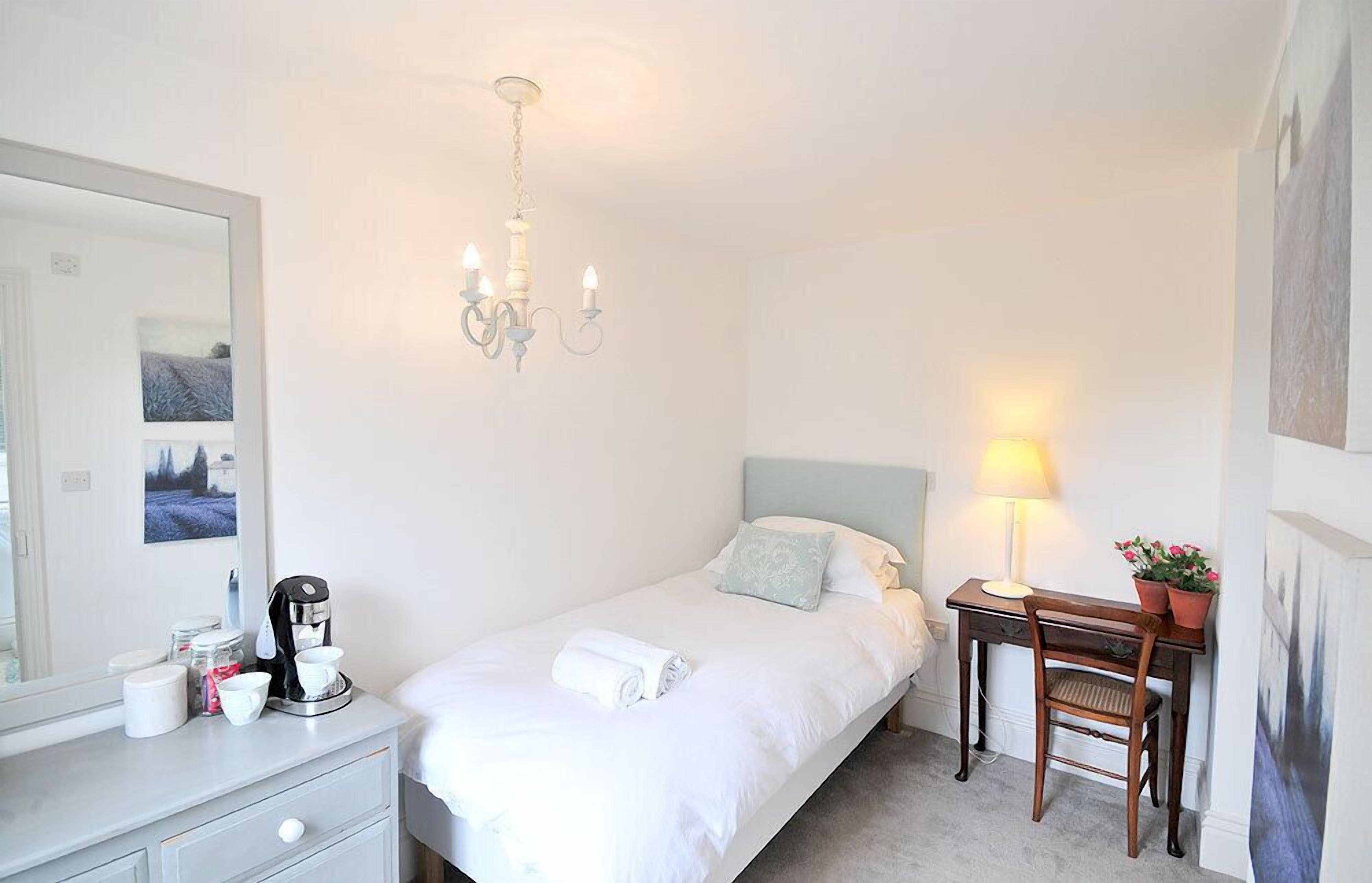 A bedroom with a bed, a desk, a chair, a lamp, a coffee maker, a mirror, and a chandelier.