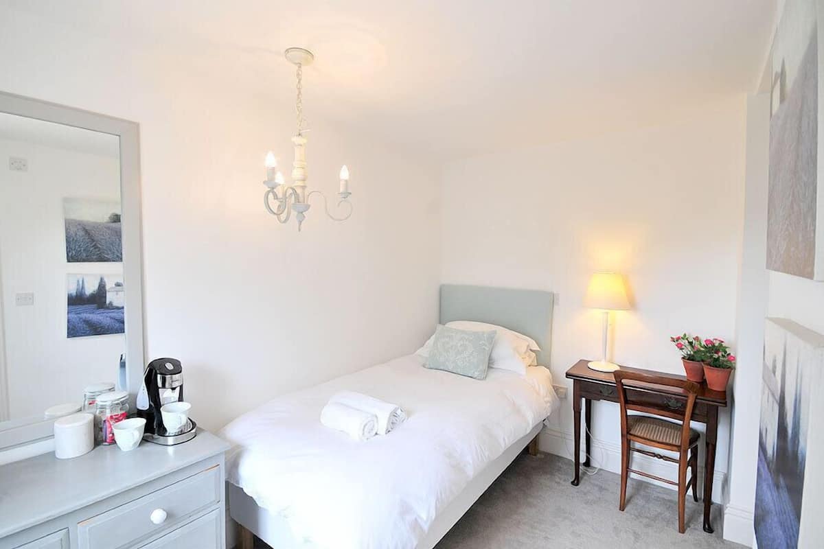 A bedroom with a bed, a desk, a chair, a lamp, a coffee maker, a mirror, and a chandelier.