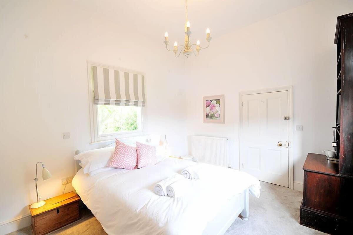 A bedroom with a bed, bedside lamp, window with blinds, radiator, and a door.