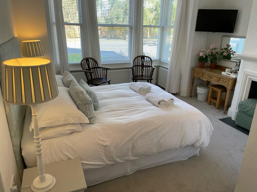 A neatly made bed with white linens, two wooden chairs, a desk with a mirror, and a television mounted on the wall.