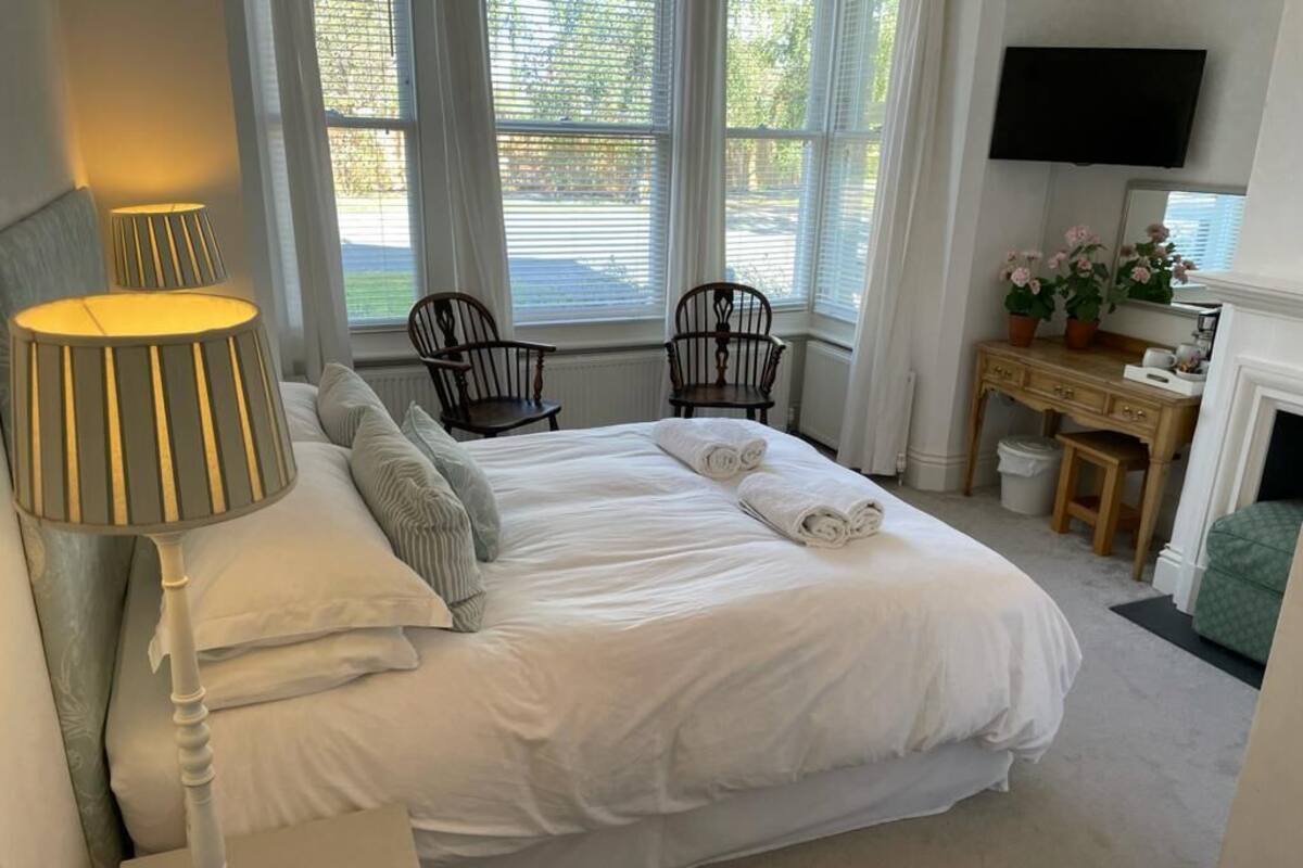 A neatly made bed with white linens, two wooden chairs, a desk with a mirror, and a television mounted on the wall.