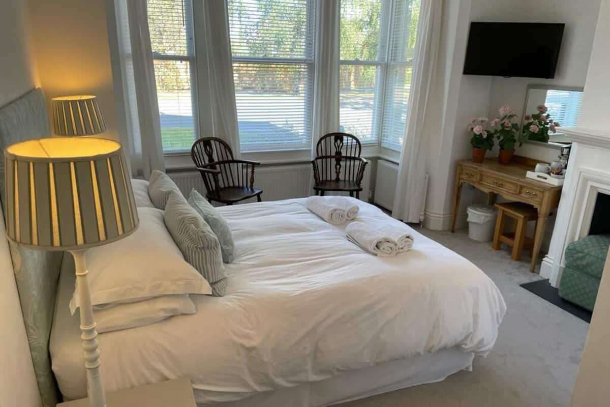 A neatly made bed with white linens, two wooden chairs, a desk with a mirror, and a television mounted on the wall.