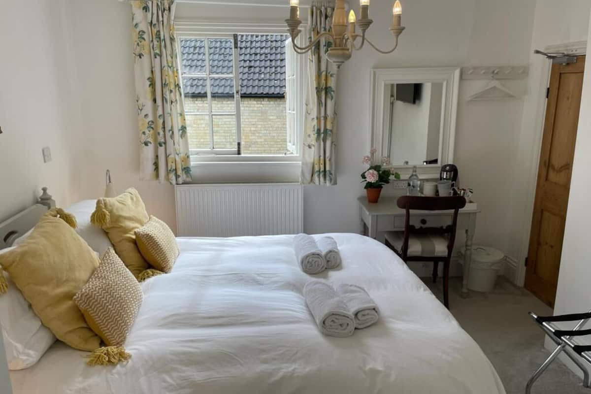 A neatly made bed with rolled towels, a bedside table, a mirror, a radiator, and a window with curtains.