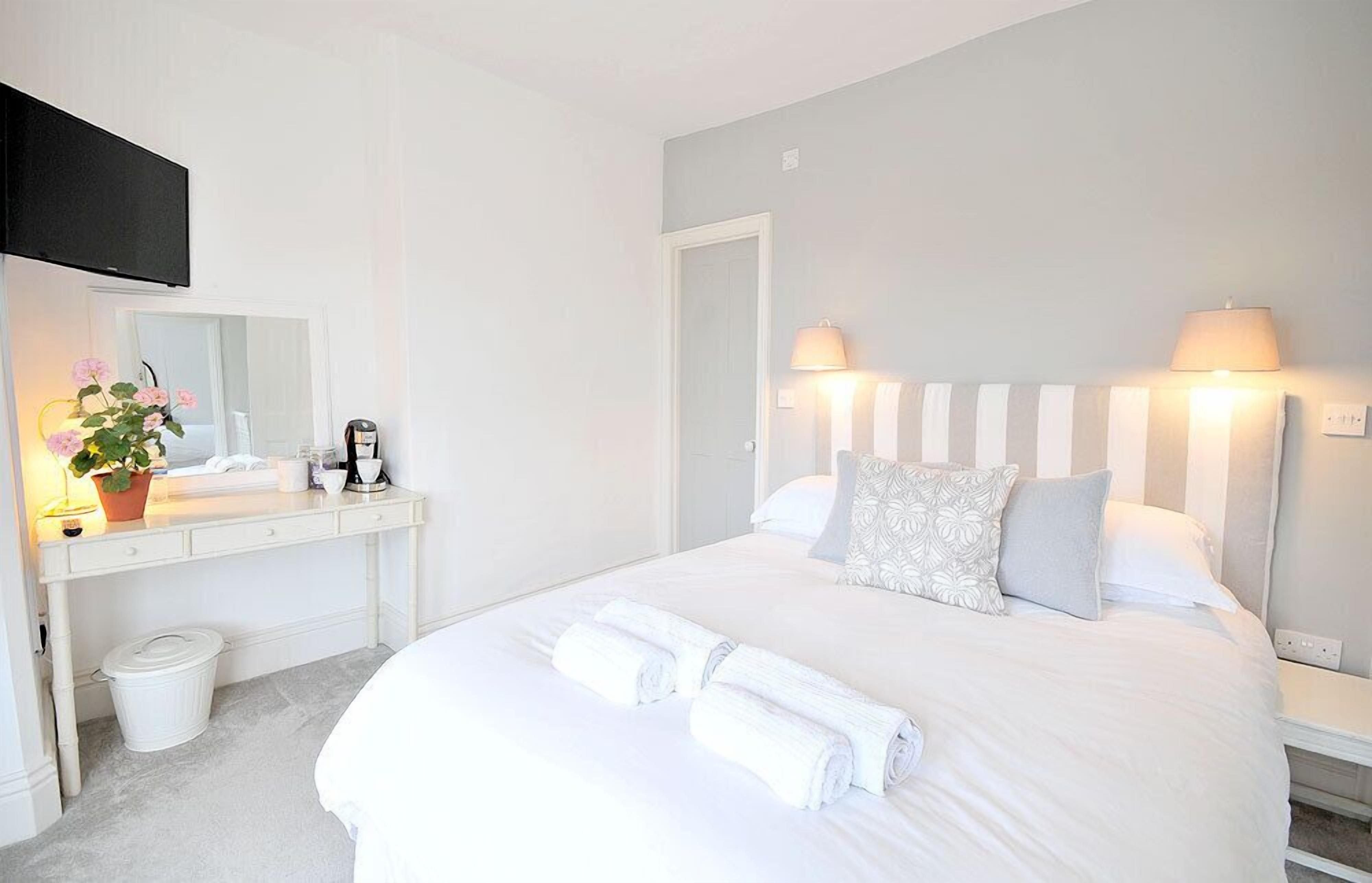 A hotel room with a bed, bedside lamps, a desk with a mirror, a trash bin, and a potted plant.