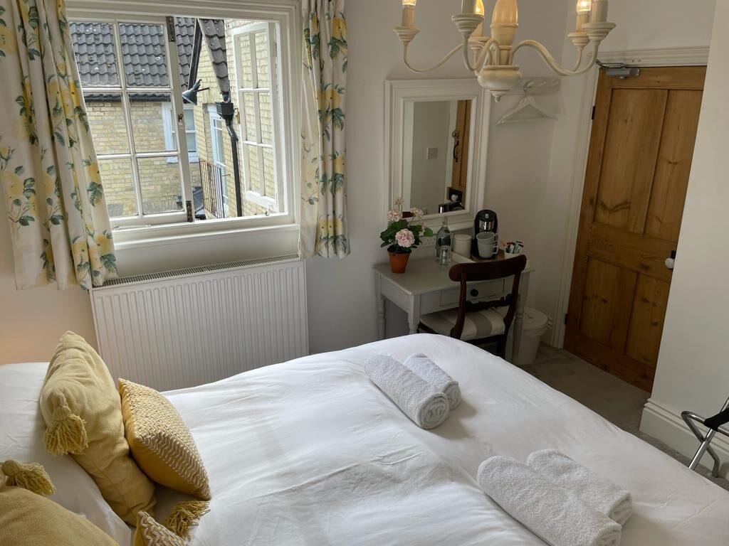 A hotel room with a bed, a window with curtains, a bedside table with a lamp, a mirror, and a wooden door.
