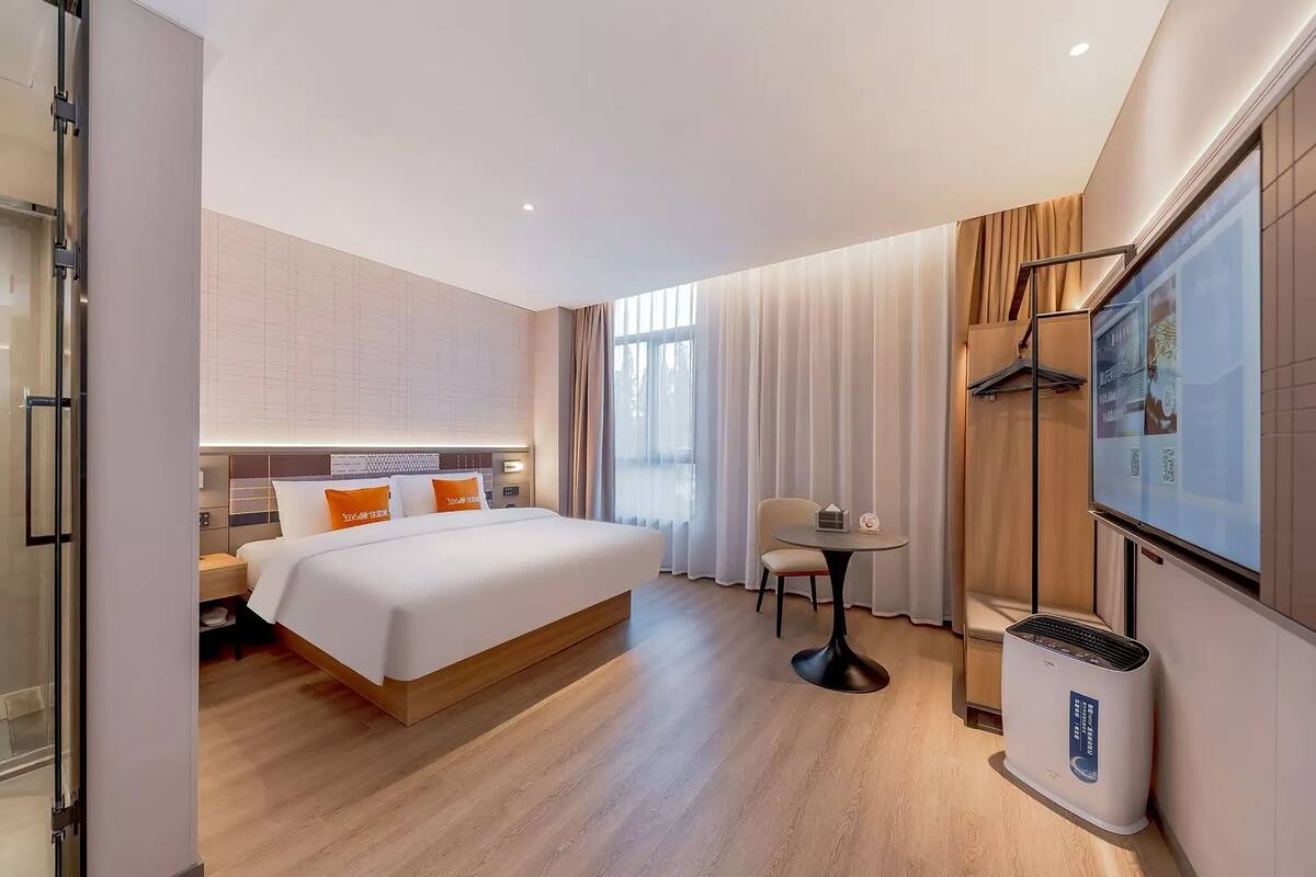 A hotel room with a bed, bedside table, chair, TV, and air conditioning unit.