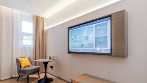 Una habitación de hotel moderna con un televisor grande integrado en la pared mostrando una interfaz de hotel, un sillón gris con un cojín amarillo, una mesita redonda pequeña y un banco con una franja naranja.