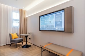 Una habitación de hotel moderna con un televisor grande integrado en la pared mostrando una interfaz de hotel, un sillón gris con un cojín amarillo, una mesita redonda pequeña y un banco con una franja naranja.
