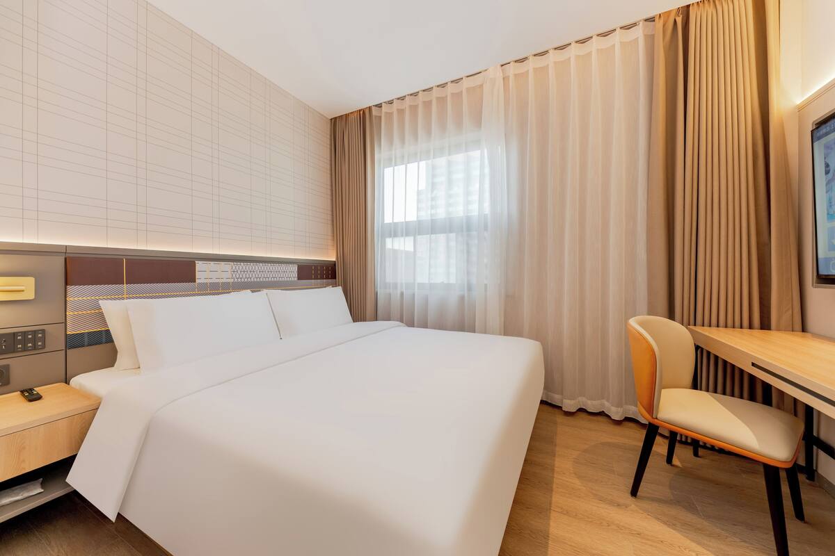 A hotel room with a large bed, a desk, a chair, a TV, and a control panel.