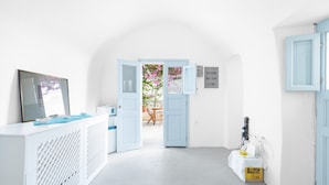 A room with a white console table, a bed with a blue headboard, a wardrobe, and an open door leading to a balcony with flowers. - Thera Bliss "Hideaway" Private Jacuzzi by MΡS (santorini)