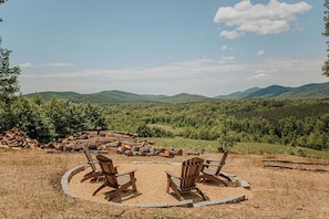 Un espacio circular para sentarse con sillas de madera y una chimenea, rodeado de un paisaje natural con colinas y árboles.